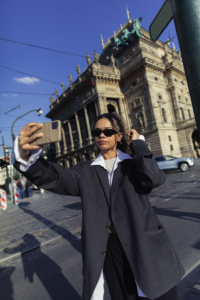 νεαρή Αφροαμερικανή με τεράστιο σακάκι και γυαλιά ηλίου που βγάζει selfie κοντά στο εθνικό θέατρο στην Πράγα - Φωτογραφία, εικόνα
