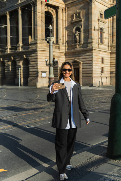 happy african american woman in oversized blazer and sunglasses taking selfie near building in prague - Photo, image