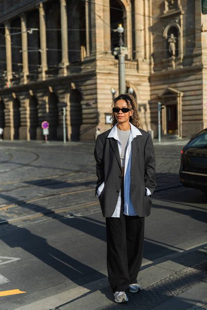 curly african american woman in oversized blazer and sunglasses walking with hands in pockets near building in prague - Foto, imagen