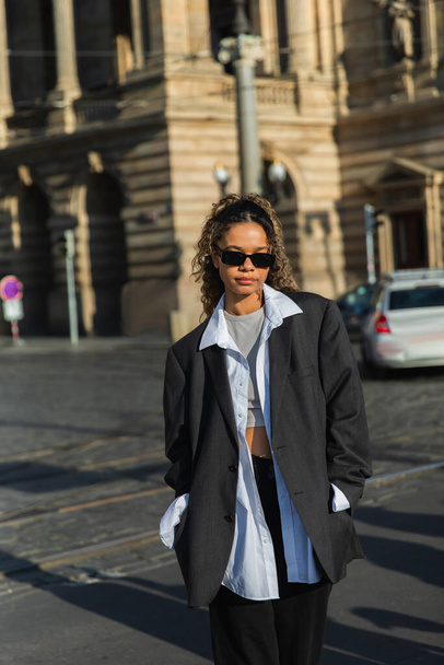 молодая африканская американка в стильном блейзере и солнцезащитных очках ходит с руками в карманах возле здания в Праге - Фото, изображение