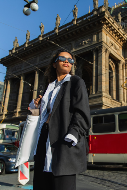 άποψη χαμηλής γωνίας της νεαρής Αφροαμερικανής γυναίκας με γυαλιά ηλίου ποζάροντας κοντά στο εθνικό θέατρο στην Πράγα - Φωτογραφία, εικόνα