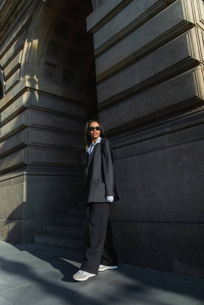 юна афроамериканська жінка в сонцезахисних окулярах і великий костюм стоять біля будинку на міській вулиці в Празі - Фото, зображення