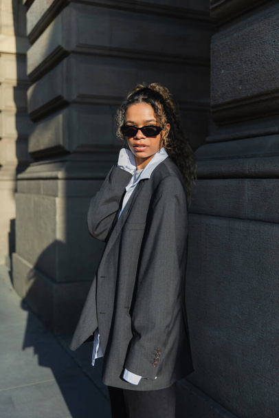 curly african american woman in sunglasses and oversize blazer walking near building on urban street in prague - Foto, Imagem