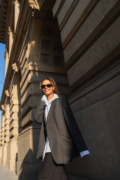 sunshine on face of happy african american woman in sunglasses and oversize suit walking on urban street in prague - Photo, image
