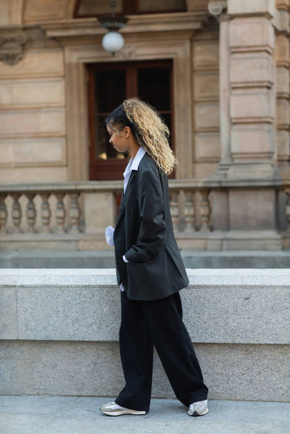 side view of displeased african american woman in oversize suit walking with hand in pocket on urban street in prague - Foto, Bild