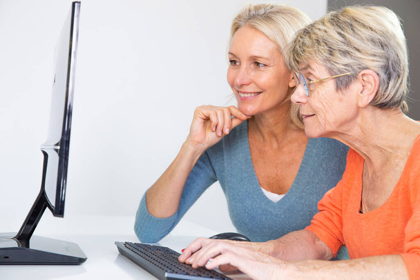 Μια πενηντάρα που βοηθάει μια ηλικιωμένη γυναίκα να χρησιμοποιήσει υπολογιστή.. - Φωτογραφία, εικόνα