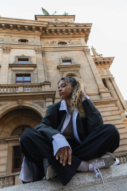 low angle view of african american woman in oversize suit sitting with crossed legs near national theatre in prague  - Fotografie, Obrázek