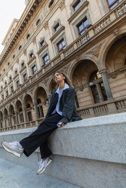 african american woman in oversize suit listening music in wireless earphone wile sitting near building in prague  - Photo, Image
