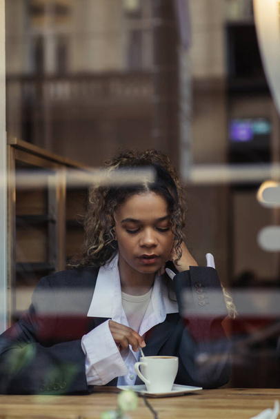 Αφροαμερικανή γυναίκα σε μοντέρνο σακάκι ανακατεύοντας καφέ στο κύπελλο, ενώ κάθεται πίσω από γυαλί παράθυρο στο καφέ - Φωτογραφία, εικόνα