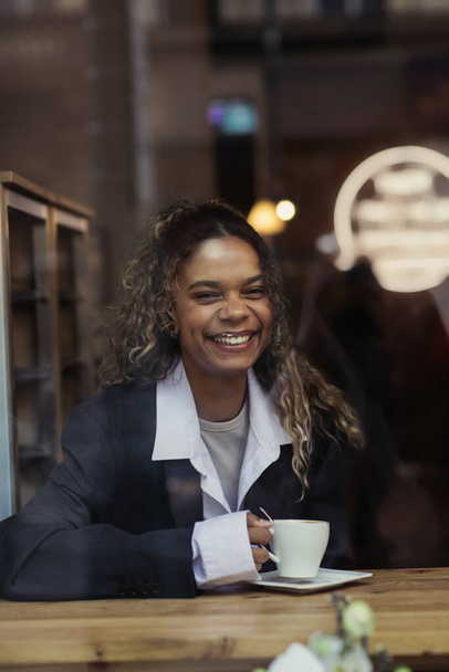 Χαρούμενη Αφροαμερικανή γυναίκα που κρατάει ένα φλιτζάνι καφέ και χαμογελά πίσω από το τζάμι του παραθύρου στο καφέ - Φωτογραφία, εικόνα