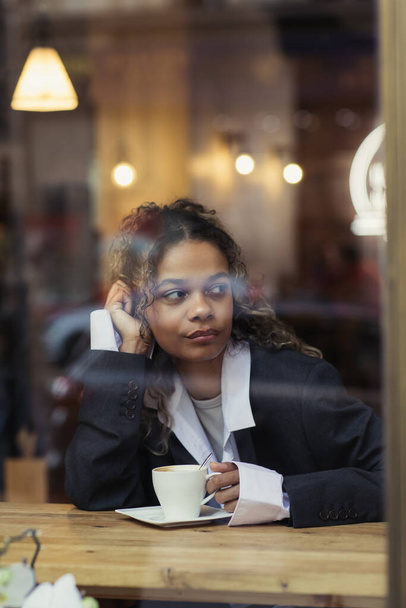pensive african american woman holding cup of coffee behind window glass in cafe - Zdjęcie, obraz