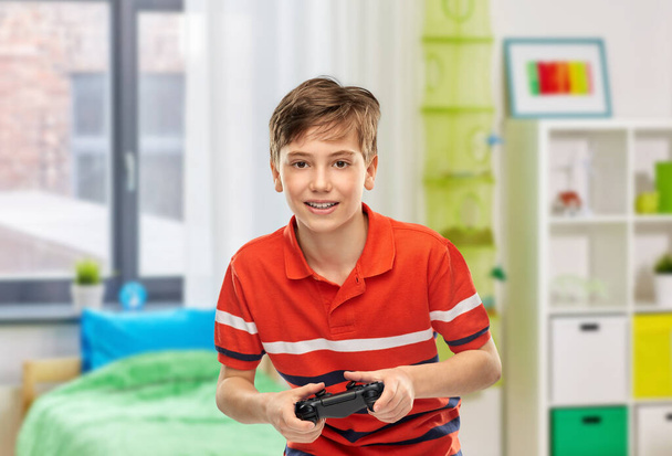 childhood, fashion and people concept - portrait of happy smiling boy in red polo t-shirt with gamepad playing video game over home room background - Foto, Imagem