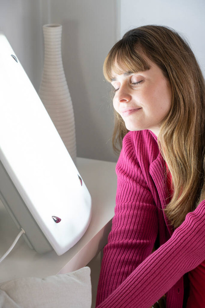 Woman sitting near a white light lamp for light therapy. - Foto, Imagen