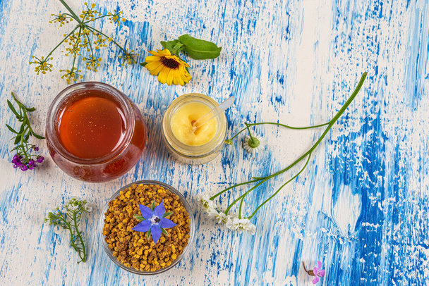 Honey, pollen and royal jelly surrounded by medicinal flowers deposited on an old board. - Photo, image