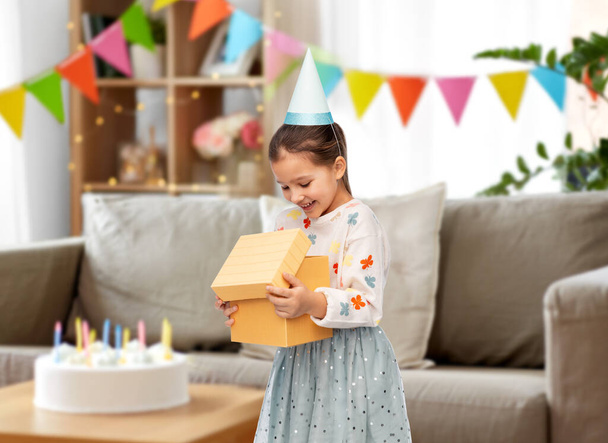 birthday, childhood and people concept - portrait of smiling little girl in party hat opening gift box over decorated home room background - Foto, afbeelding