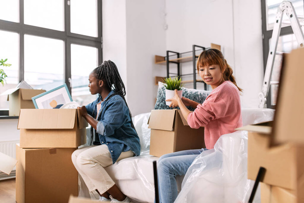 moving, people and real estate concept - happy smiling women unpacking boxes at new home - 写真・画像