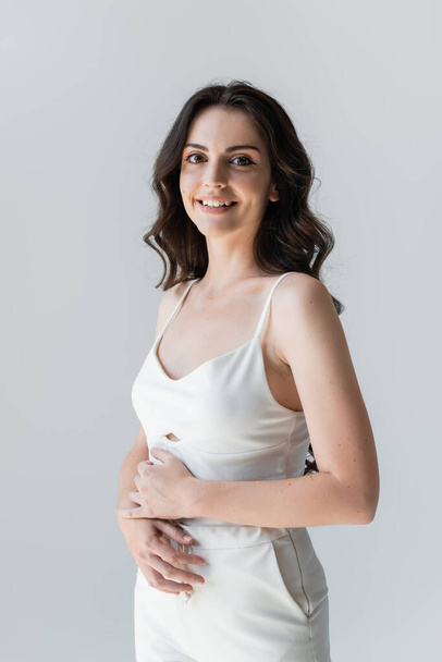 Portrait of smiling brunette woman in white clothes looking at camera isolated on grey  - Fotografie, Obrázek