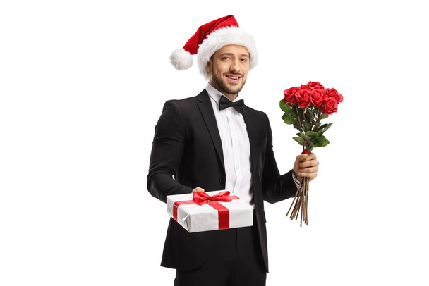 Handsome man in a suit and bow tie wearing a santa claus hat and holding roses and gift box isolated on white background - Fotoğraf, Görsel