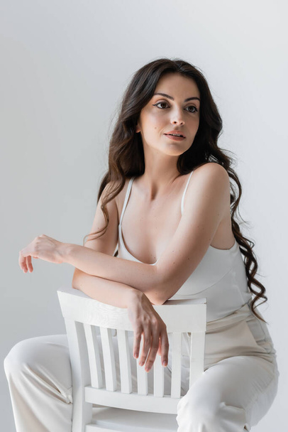 Brunette woman in white clothes sitting on chair isolated on grey  - Foto, Imagen