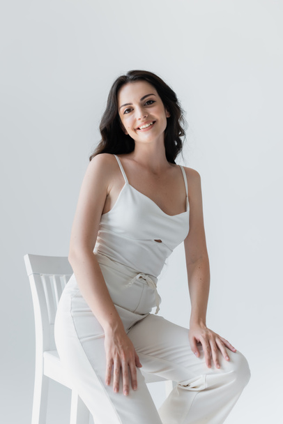 Pretty woman in white clothes smiling near chair isolated on grey  - Фото, зображення