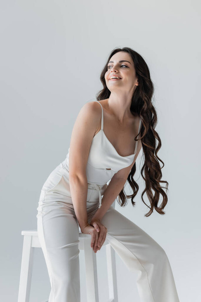 Positive long haired model sitting on chair and looking away isolated on grey  - Foto, Bild