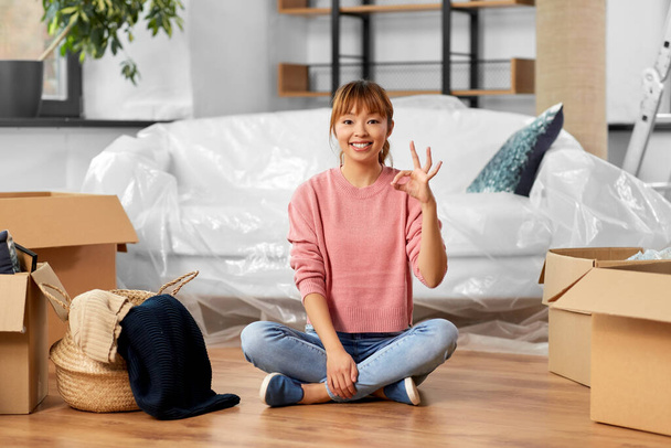moving, people and real estate concept - happy smiling woman with boxes sitting on floor and showing ok sing at new home - Φωτογραφία, εικόνα