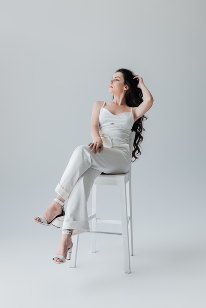 Full length of young model in white clothes and heels sitting on chair on grey background - Foto, Imagen