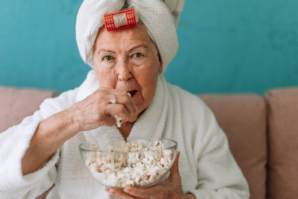 Happy senior couple sitting at a sofa in bathrobes and watching TV with popcorn. - 写真・画像