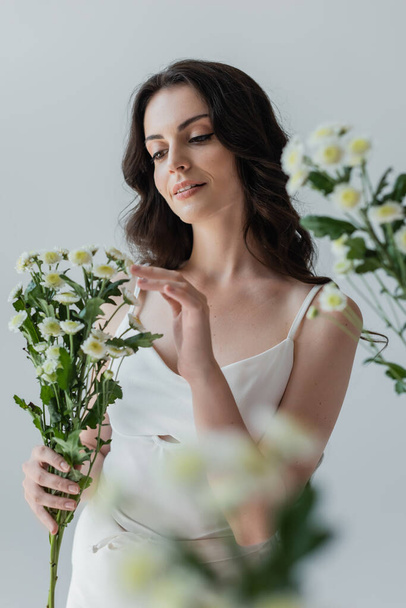 Pretty brunette woman in white top touching flowers isolated on grey  - Foto, Imagem