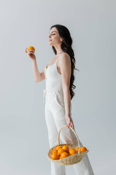 Smiling woman in white clothes holding basket with fresh oranges isolated on grey  - Foto, immagini