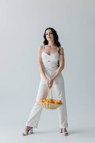 Full length of trendy woman holding basket with oranges on grey background - 写真・画像