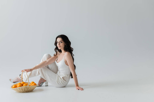 Pretty and stylish woman sitting near wicker basket with oranges on grey background - 写真・画像