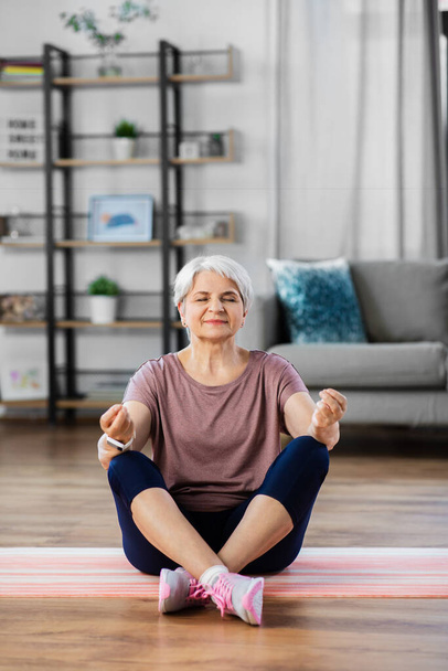 sport, fitness and healthy lifestyle concept - happy senior woman meditating on exercise mat at home - Fotoğraf, Görsel