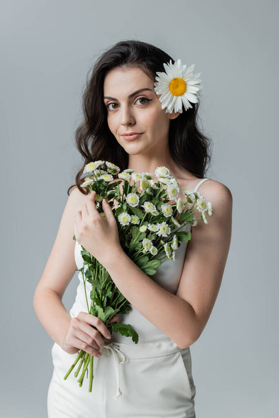 Pretty young woman holding bouquet of chamomiles while standing isolated on grey  - Foto, immagini