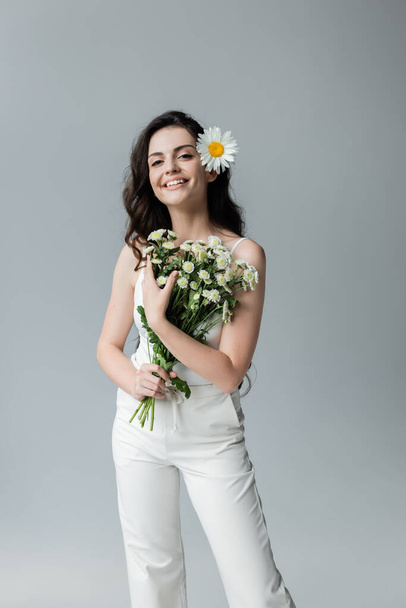Positive woman in white clothes holding bouquet of chamomiles isolated on grey  - Photo, image