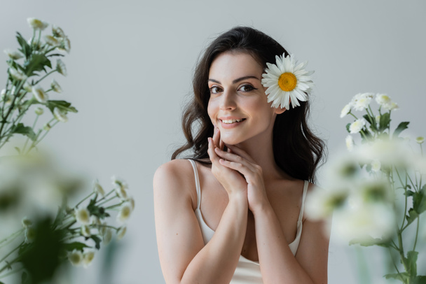 Pretty brunette woman with flower in hair smiling at camera near blurred flowers isolated on grey  - Foto, afbeelding