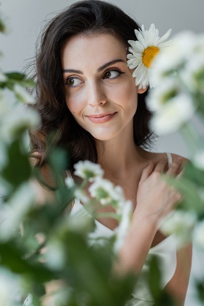 Smiling woman with visage and chamomile in hair looking away near blurred plants isolated on grey  - Valokuva, kuva