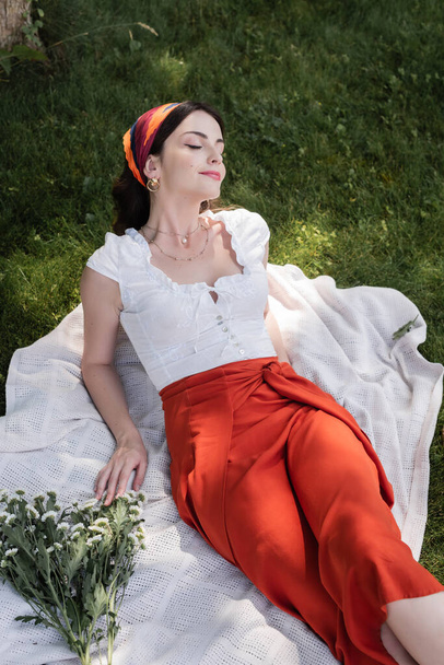 High angle view of stylish woman in blouse resting on blanket near flowers in park  - Foto, afbeelding