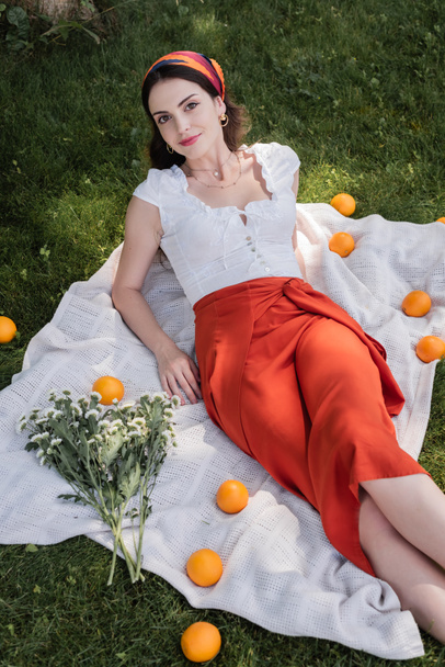 High angle view of stylish woman looking at camera near flowers and oranges on blanket in park  - Фото, зображення