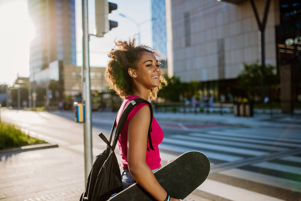 Multiracial teenage girl with backpack and skateboard, walking in city during a summer day. - Фото, изображение