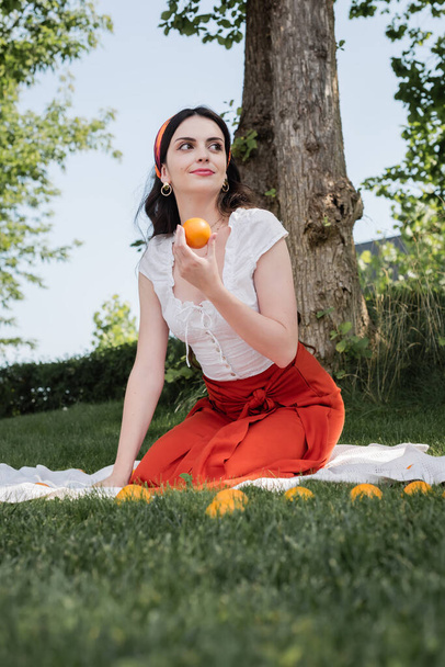 Brunette woman in blouse and skirt holding oranges on blanket in park  - Foto, afbeelding
