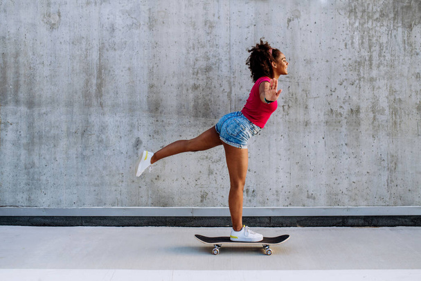 Multiracial teenage girl riding skateboard in front of concrete wall, standing in one leg, balancing. Side view. - Фото, зображення