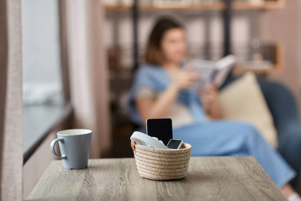 digital detox and leisure concept - close up of gadgets in basket on table and woman reading book at home - Фото, зображення
