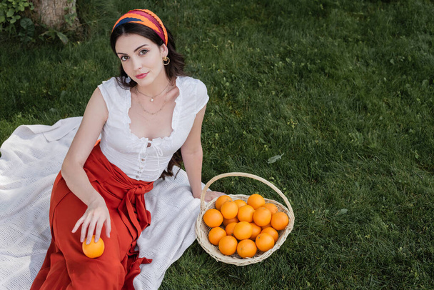 High angle view of smiling woman in blouse holding orange while sitting on blanket in park  - Φωτογραφία, εικόνα