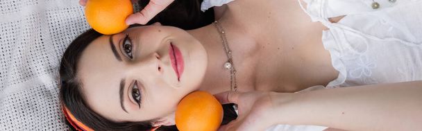 Top view of brunette woman in blouse holding oranges on blanket outdoors, banner  - Foto, immagini