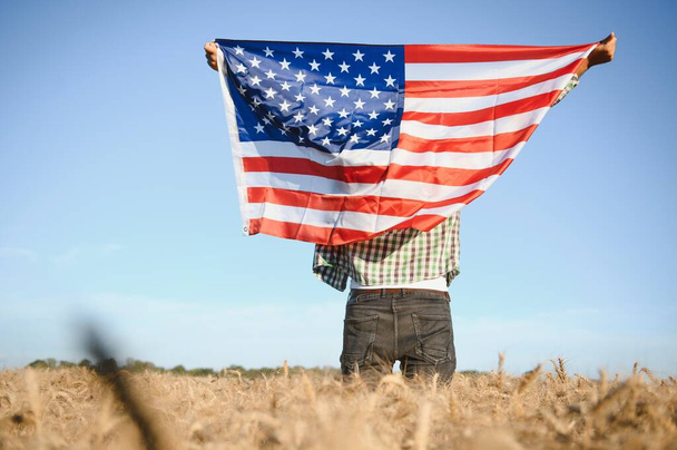 Young patriotic farmer stands among new harvest. Boy walking with the american flag on the wheat field celebrating national independence day. 4th of July concept - 写真・画像
