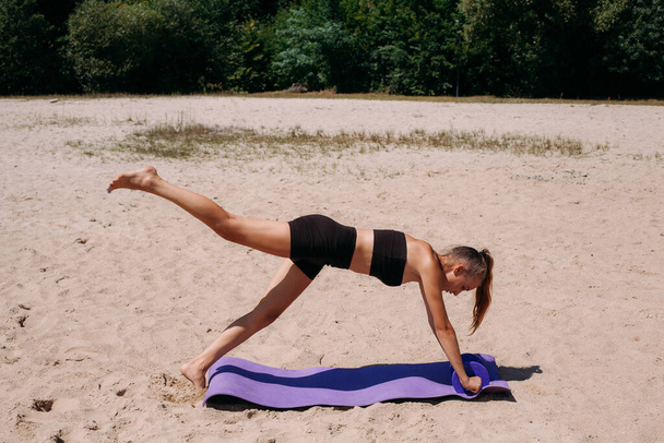 A beautiful tanned girl with a slim figure and blond hair, in black shorts and a tank top, is doing fitness with dumbbells, on a mat, on the beach in summer. - Fotografie, Obrázek
