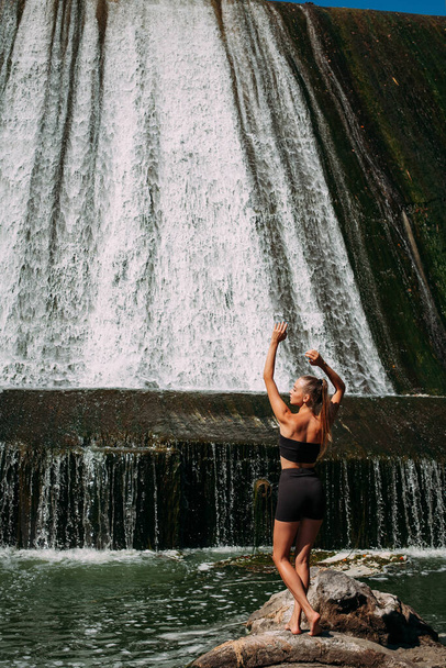 A slender tanned girl with a sports figure is doing yoga exercises against the backdrop of a picturesque waterfall in the summer. She is wearing black shorts and a top. - Fotó, kép