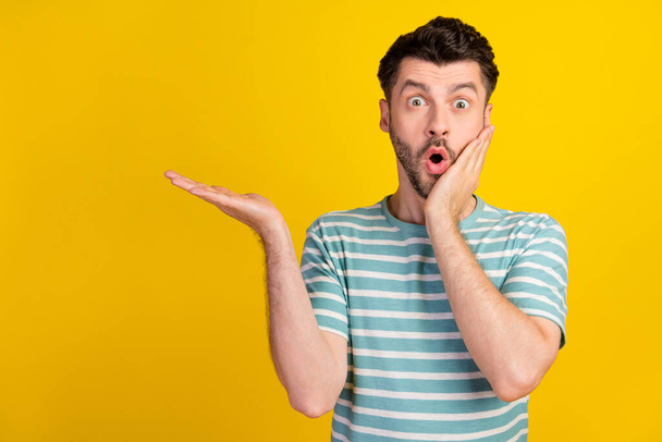 Photo of ecstatic impressed man dressed striped t-shirt demonstrating empty space hand on cheek isolated on yellow color background. - Foto, imagen