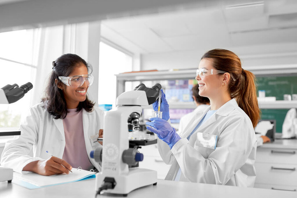 science research, work and people concept - happy international female scientists with microscopes working in laboratory - Photo, image